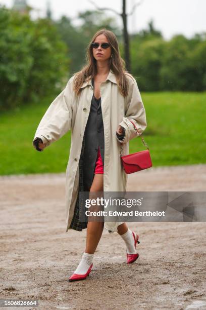 Guest wears black vintage sunglasses, a dark gray buttoned long gilet, a red shorts, a black V-neck tank-top, a beige long coat, a red shiny leather...