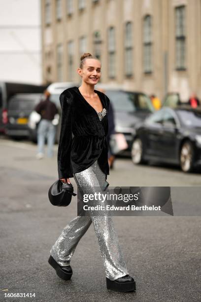 Guest wears a black V-neck belted velvet jacket, a silver embroidered sequined V-neck / flared jumpsuit, a black shiny leather handbag, black shiny...