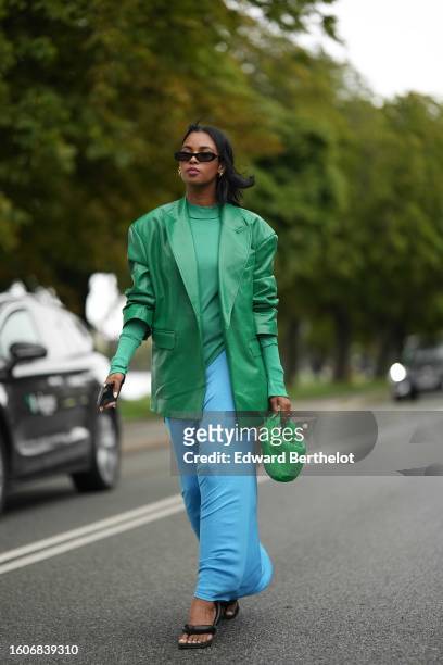Guest wears black sunglasses, gold earrings, a green turtleneck wool asymmetric top, a flashy green shiny leather blazer jacket, a flashy blue long...