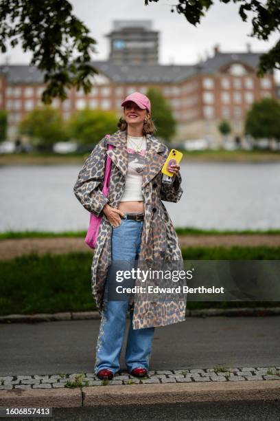 Guest wears a pink denim cap, silver earrings, gold chain pendant necklaces, a white with black cat print pattern cropped t-shirt, a beige / brown /...