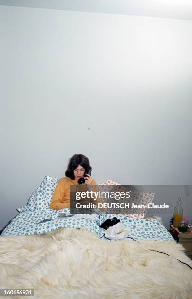 Close-up Of Antoine. ANTOINE, asssis au fond de son lit, vêtu d'un polo jaune téléphone avec un combiné noir posé sur une parure de draps à fleurs...