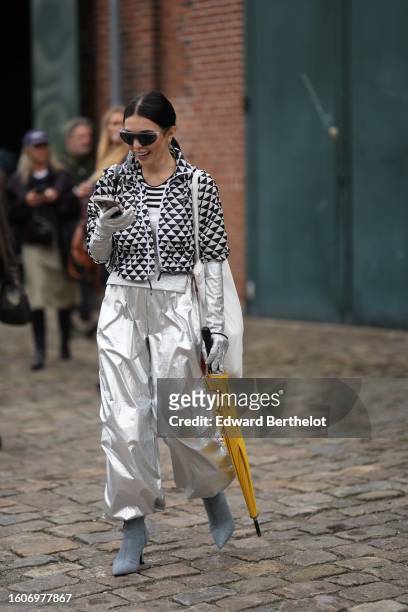 Guest wears black futurist sunglasses, silver small earrings, a black and white triangular print pattern zipper jacket from Prada, silver shiny...