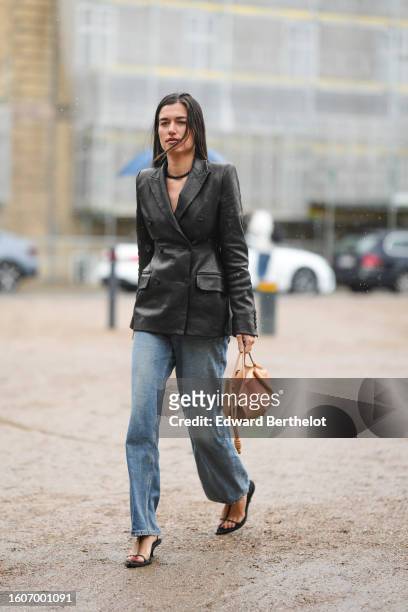 Guest wears a black necklace, a black shiny leather jacket, blue and yellow faded denim large pants, a beige brown shiny leather handbag, black...