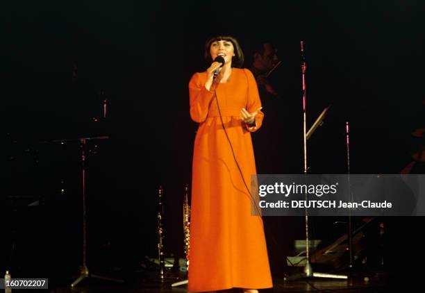 Close-up Of Mireille Mathieu In Gdr . RDA- 1972- Mireille MATHIEU, vêtue d'une longue robe orange à manches longues, chante debout sur scène, micro...