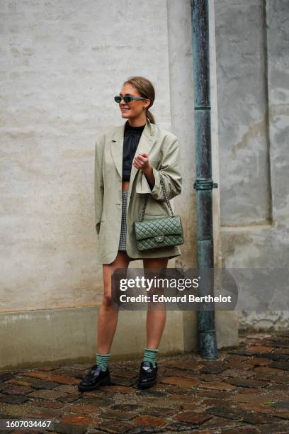 Guest wears gray sunglasses, blue stones earrings, a black high neck cropped silk top, a pale green oversized blazer jacket, a pale gray short skirt,...