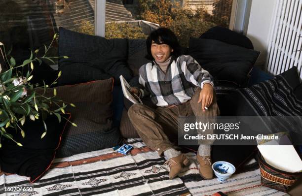 Close-up Of Kenzo Takada. En novembre 1975, dans une pièce, devant une baie vitrée, assis sur des coussins, à côté d'une plante, le styliste KENZO...