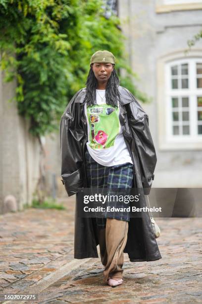 Guest wears a khaki beret from Kangol, a silver chain logo necklace from Chanel, a white with green and pink print pattern t-shirt, a black shiny...