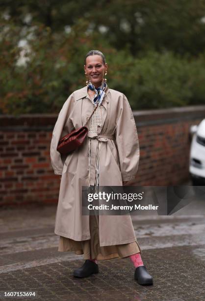 Guest seen outside Skall Studio show wearing golden earrings, blue Chanel silk scarf, crocheted light yellow top, beige oversized trenchcoat,...