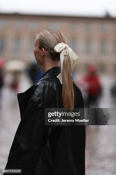 Guest seen wearing silver earrings, cream white satin and silk ribbon as hair accessory, black leather long coat, outside The Garment, during the...