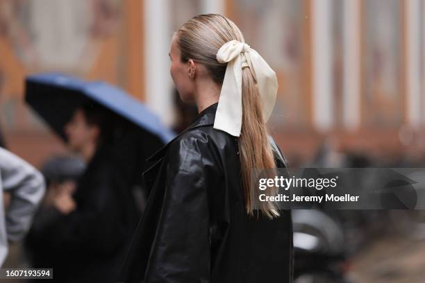 Guest seen wearing silver earrings, cream white satin and silk ribbon as hair accessory, black leather long coat, outside The Garment, during the...