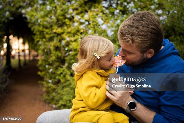 father and daughter eating candy, lollipop together sitting on bench in park in nature,copy space - lollipop man stockfoto's en -beelden