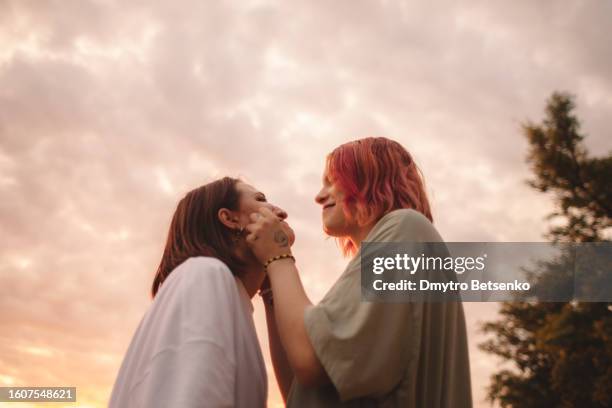 happy young woman touching her girlfriends cheeks at sunset - lesbica foto e immagini stock