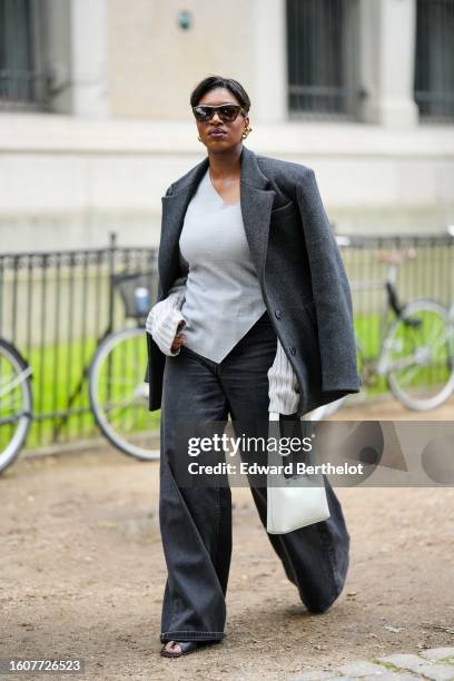 Loicka Grace wears dark brown sunglasses, gold earrings, a dark gray felt wool coat, a pale gray linen blouse, dark gray denim large wide legs pants,...