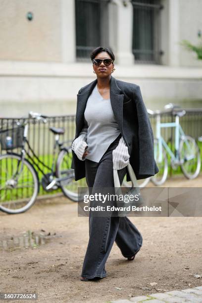 Loicka Grace wears dark brown sunglasses, gold earrings, a dark gray felt wool coat, a pale gray linen blouse, dark gray denim large wide legs pants,...