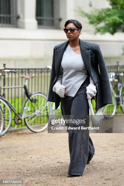 Loicka Grace wears dark brown sunglasses, gold earrings, a dark gray felt wool coat, a pale gray linen blouse, dark gray denim large wide legs pants,...