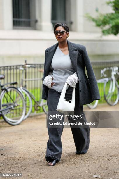 Loicka Grace wears dark brown sunglasses, gold earrings, a dark gray felt wool coat, a pale gray linen blouse, dark gray denim large wide legs pants,...