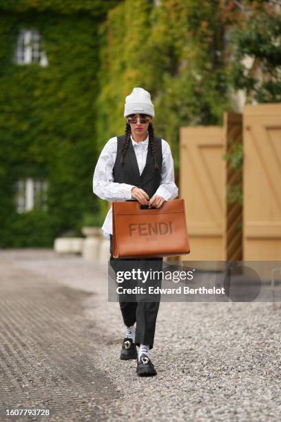 Katya Tolstova wears a white wool beanie from Munthe, brown sunglasses, silver earrings, a white with small black print pattern shirt, a black with...