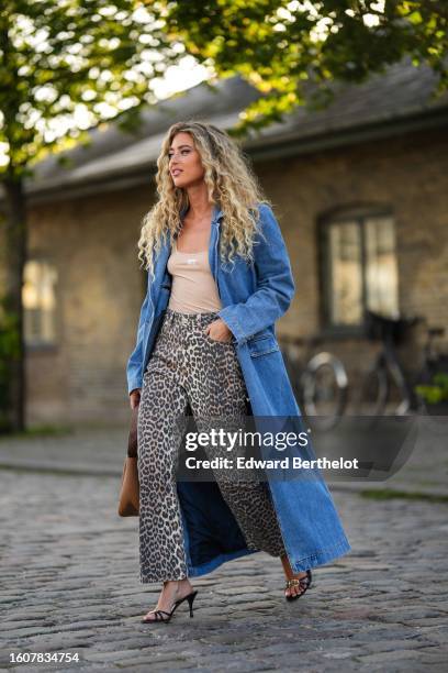Emili Sindlev wears a blue denim long coat, a beige tank-top from Miu Miu, beige and black leopard print pattern denim flared pants, a camel shiny...