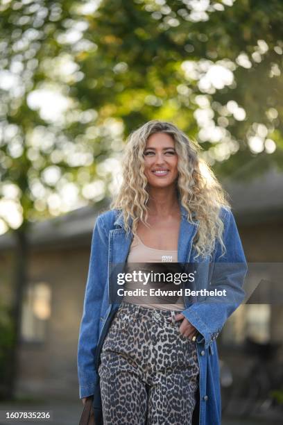 Emili Sindlev wears a blue denim long coat, a beige tank-top from Miu Miu, beige and black leopard print pattern denim flared pants, a camel shiny...