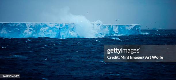 a large iceberg with steep sides, floating on the water. waves and spray rising. calving event. - glacial ice sheet stock pictures, royalty-free photos & images