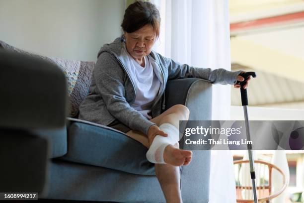 a sad senior asian woman sitting on sofa holding a walking stick with a bandage on her foot - twisted ankle stock pictures, royalty-free photos & images