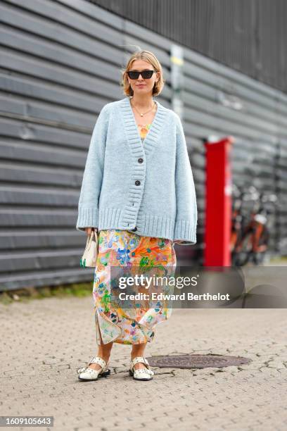 Guest wears black sunglasses from Saint Laurent, a gold chain pendant necklace, a pale blue oversized wool pullover cardigan, a white with orange /...