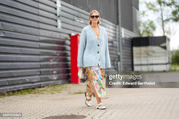 Guest wears black sunglasses from Saint Laurent, a gold chain pendant necklace, a pale blue oversized wool pullover cardigan, a white with orange /...