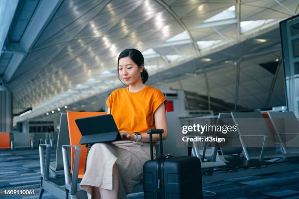 young asian businesswoman working with digital tablet while waiting for her flight in airport terminal, a suitcase by her side. young asian woman on business trip. lifestyle and technology. travel and vacation concept. business on the go - business class imagens e fotografias de stock