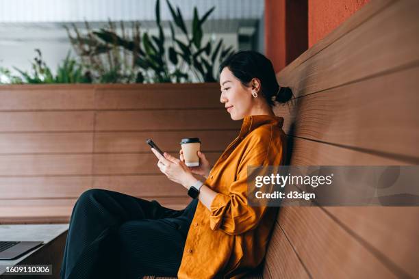 young asian woman sitting in a sidewalk cafe with a cup of coffee and using her smartphone. enjoying a relaxing moment in the afternoon. coffee break. lifestyle and technology - telefoon gebruiken stockfoto's en -beelden