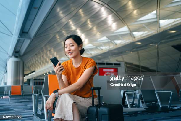 young asian woman with suitcase using smartphone while waiting for her flight at airport terminal. asian businesswoman on business travel. lifestyle and technology. travel and vacation concept - travel fotografías e imágenes de stock