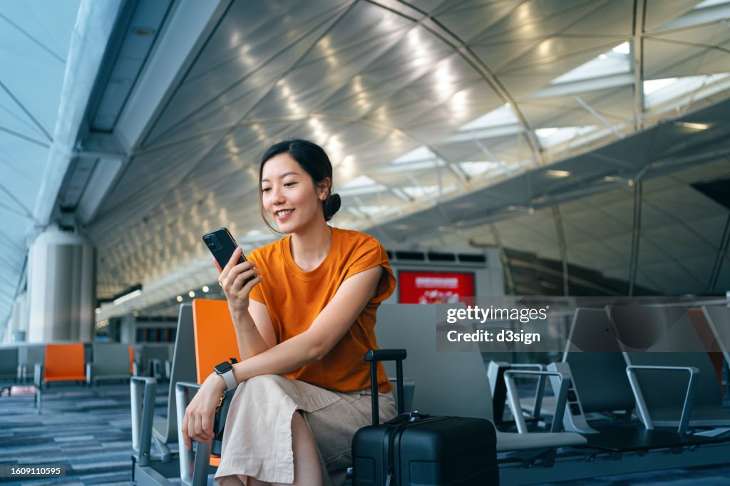 Young Asian woman with suitcase using smartphone while waiting for her flight at airport terminal. Asian businesswoman on business travel. Lifestyle and technology. Travel and vacation concept
