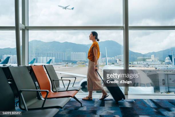 young asian woman carrying suitcase, walking by the window at airport terminal. young asian female traveller waiting for boarding at airport. business travel. travel and vacation concept - travel stock-fotos und bilder