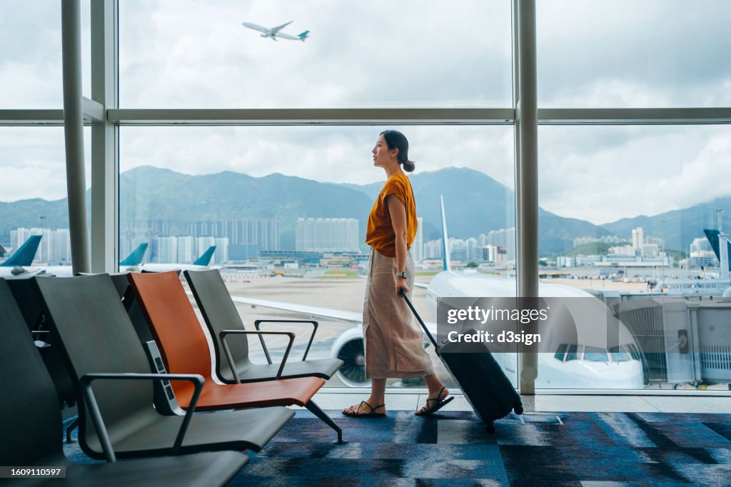 Young Asian woman carrying suitcase, walking by the window at airport terminal. Young Asian female traveller waiting for boarding at airport. Business travel. Travel and vacation concept