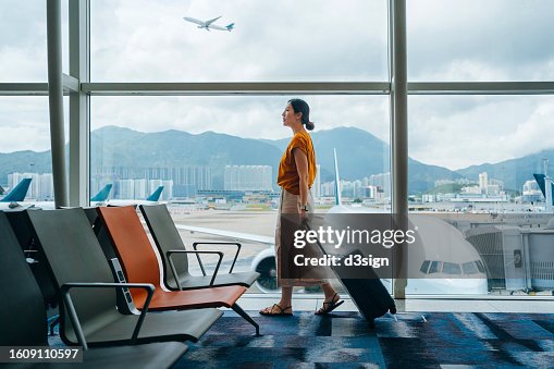 Young Asian woman carrying suitcase, walking by the window at airport terminal. Young Asian female traveller waiting for boarding at airport. Business travel. Travel and vacation concept