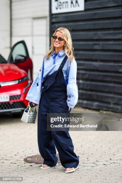 Guest wears black sunglasses, a pale blue puffy sleeves shirt, navy blue denim large overalls, a silver braided shiny leather handbag, white heels...