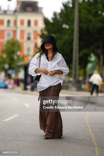 Guest wears black large oversized bob hat, a white shirt, a black shiny leather shoulder bag, diamonds rings, a pale yellow silk flower brooch, a...