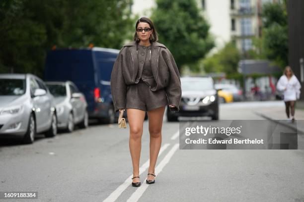 Guest wears black small sunglasses, a dark brown wool buttoned gilet, a matching dark brown zipper oversized bomber coat, dark brown wool shorts, a...