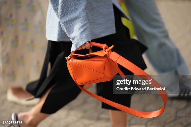 Guest wears a blue gray shoulder-pad / belted blazer jacket, a black large fringed midi skirt, a neon orange shiny leather Puzzle handbag from Loewe,...