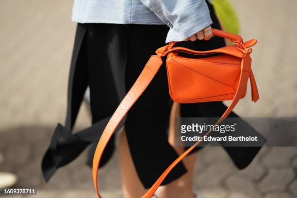 Guest wears a blue gray shoulder-pad / belted blazer jacket, a black large fringed midi skirt, a neon orange shiny leather Puzzle handbag from Loewe,...