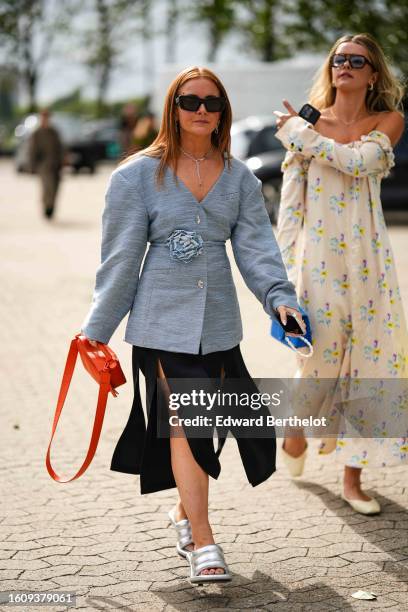 Guest wears black sunglasses, a silver large chain pendant necklace, a blue gray shoulder-pad / belted blazer jacket, a black large fringed midi...