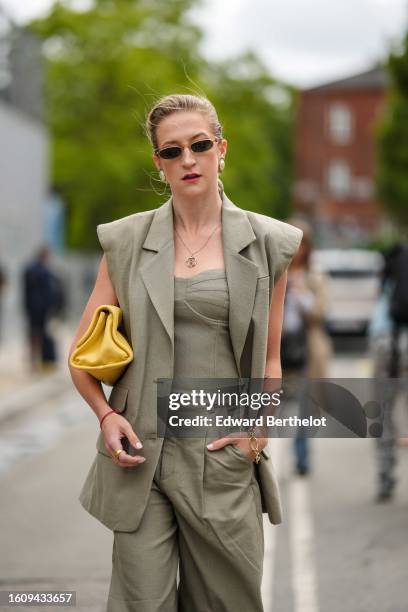 Guest wears black sunglasses, a gold chain pendant necklace, a pale khaki sleeveless / shoulder-pads gilet, matching pale khaki flared suit pants, a...