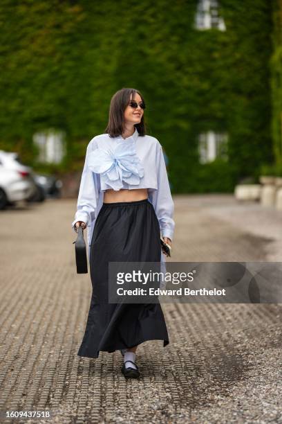 Guest wears black sunglasses, silver earrings, a pale blue high neck / cropped shirt with a large embroidered flower pattern, a black high waist long...