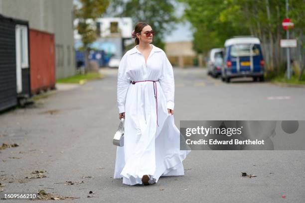 Guest wears a pale pink satin hair elastic, brown sunglasses, gold earrings, a white V-neck / puffy long sleeves oversized long shirt dress, a purple...