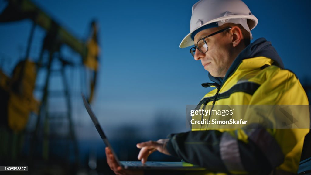 Serious engineer using laptop while working at oil field against sky during dusk