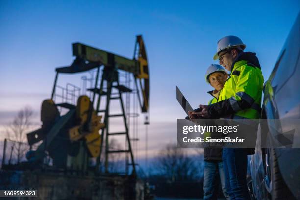 partner che pianificano su laptop mentre si trovano contro le attrezzature in auto sul campo - piattaforma petrolifera foto e immagini stock