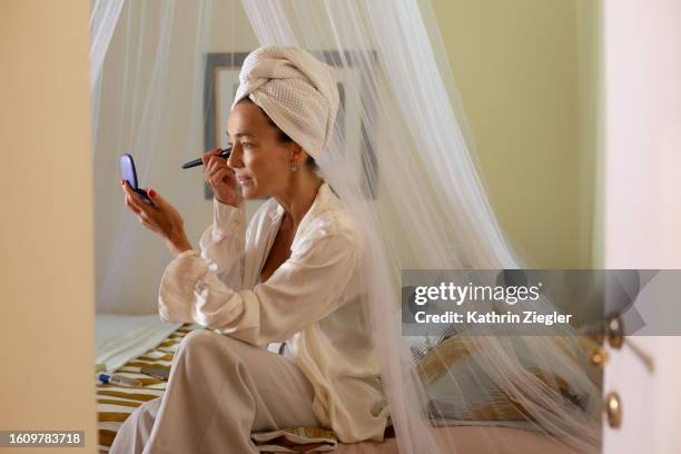woman sitting on bed, applying make-up with brush - cream colored shirt stock-fotos und bilder
