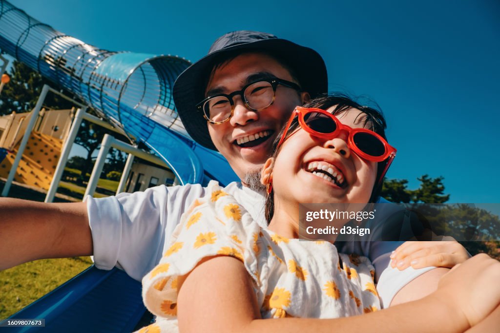 Happy young Asian father with her little daughter having fun playing on the tube slide together in an outdoor playground on a lovely sunny day. Outdoor fun. Happy childhood and fatherhood. Father and daughter bonding moment. Happy family life
