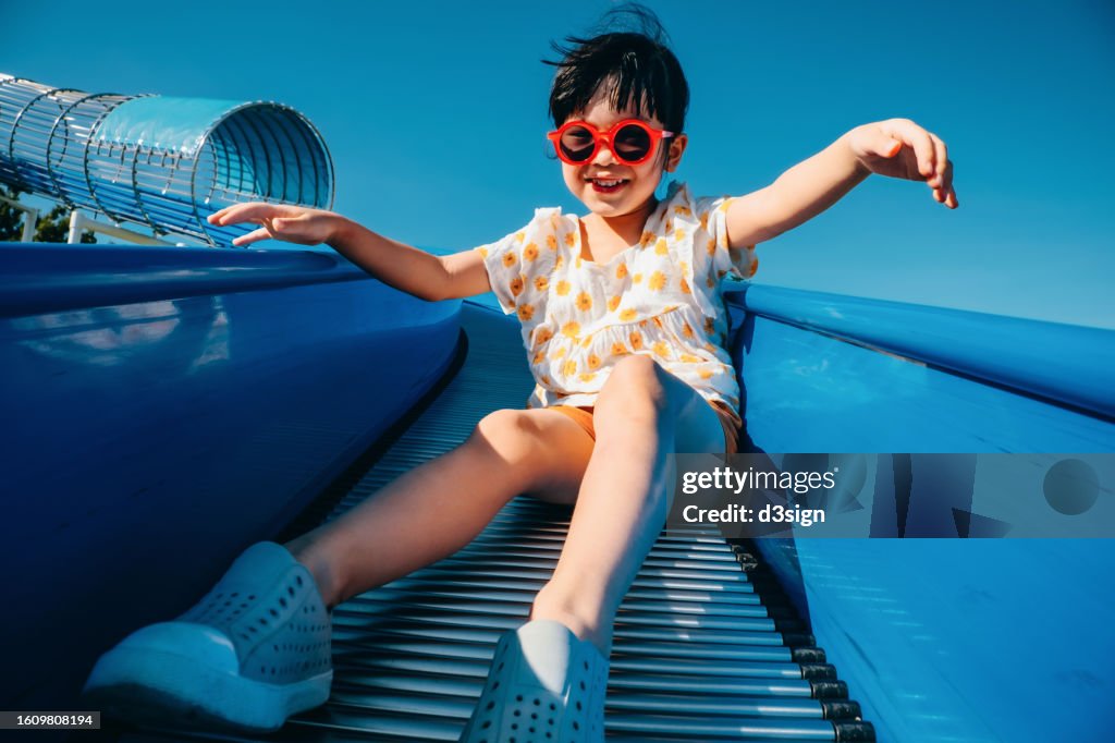 Joyful little Asian girl having fun playing on the tube slide in an outdoor playground on a lovely sunny day. Outdoor fun. Carefree and freedom concept