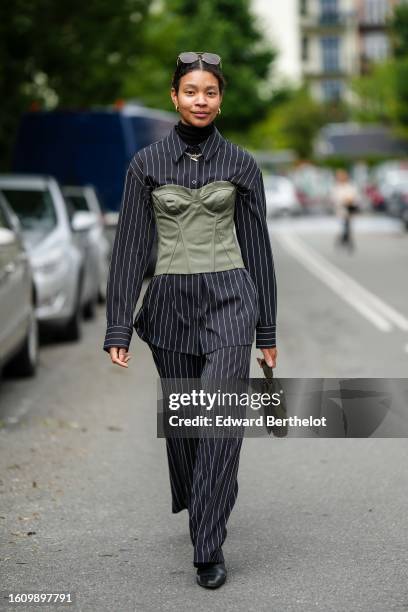 Guest wears brown sunglasses, a black turtleneck pullover, gold necklaces, a black striped print pattern blazer jacket, matching black striped print...