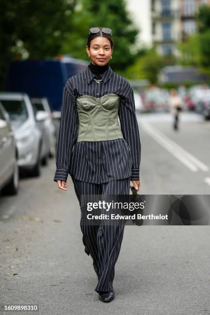 Guest wears brown sunglasses, a black turtleneck pullover, gold necklaces, a black striped print pattern blazer jacket, matching black striped print...